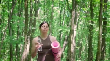 Spor yoga minderiyle çekici atletik kadın şişeden su içiyor ve yaz parkında sabah sporundan sonra dışarıda yürüyor, sağlıklı yaşam tarzı ve rehidrasyon kavramı.