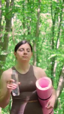 Spor yoga minderli çekici bir kadın şişeden su içiyor ve yaz parkında sabah sporundan sonra dışarıda yürüyor, sağlıklı yaşam tarzı ve rehidrasyon kavramı, dikey