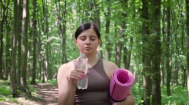 Spor yoga minderiyle çekici atletik kadın şişeden su içiyor ve yaz parkında sabah sporundan sonra dışarıda yürüyor, sağlıklı yaşam tarzı ve rehidrasyon kavramı.