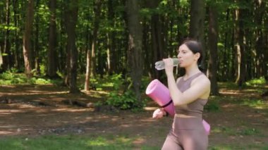 Spor yoga minderiyle çekici atletik kadın şişeden su içiyor ve yaz parkında sabah sporundan sonra dışarıda yürüyor, sağlıklı yaşam tarzı ve rehidrasyon kavramı.