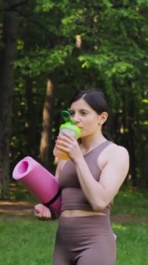 Yoga minderli çekici atletik bir kadın bcaa amino asitlerle spor besleyici içiyor ve bir salıncaktan vitaminler alıyor, sabah sporundan sonra bir yaz parkında açık havada, sağlıklı yaşam tarzı konsepti, dikey