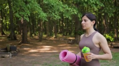 Yoga minderli sportif bir kadın bcaa amino asitlerle spor besleyici içiyor ve Shaker 'dan vitaminler alıyor, yaz parkında sabah antrenmanı, yavaş çekim, sağlıklı yaşam tarzı konsepti.