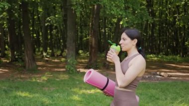 Yoga minderli sportif bir kadın bcaa amino asitlerle spor besleyici içiyor ve Shaker 'dan vitaminler alıyor, yaz parkında sabah antrenmanı, yavaş çekim, sağlıklı yaşam tarzı konsepti.