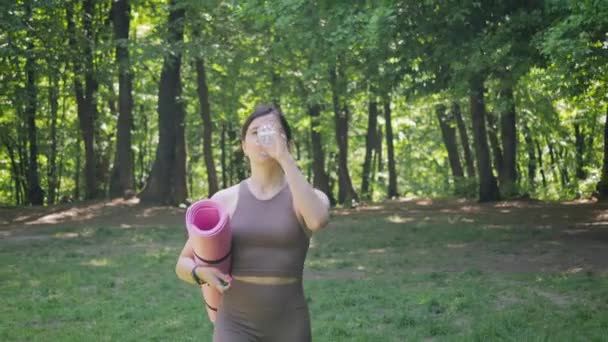 スポーツヨガマットを持つ魅力的な運動女性は ボトルから水を飲み 朝のワークアウト 健康的なライフスタイルと水分補給の概念の後に夏の公園で屋外を歩く — ストック動画