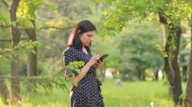 Siyah elbiseli neşeli, şık genç bayan güneşli yaz parkında açık havada akıllı telefon kullanıyor.