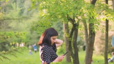 Siyah elbiseli neşeli, şık genç bayan güneşli yaz parkında açık havada akıllı telefon kullanıyor.
