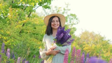 Güneşli yaz mevsiminde tarlada yürüyen, çiçekli bir buket şapkalı ve elbiseli, gülümseyen çekici genç bir kadın.