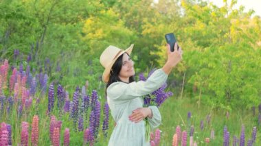 Gülümseyen çekici bir genç kadın elbise ve buket lupin çiçekli bir şapka ile güneşli yaz tarlasında akıllı bir telefondan selfie çekiyor.