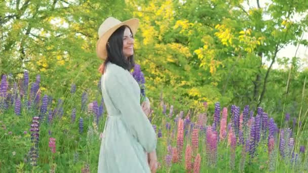 ドレスを着た笑顔の魅力的な若い女性と花束のルパンの花を保持し 日当たりの良い夏のフィールドで楽しみを持っている帽子 — ストック動画