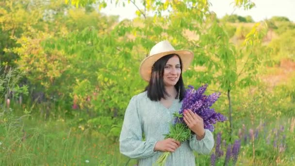 一位笑容可亲的年轻女子 身穿连衣裙 头戴礼帽 捧着鲜花 在夏日阳光灿烂的田野里尽情欢乐 — 图库视频影像