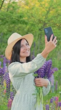 Gülümseyen çekici bir genç kadın elbise ve buket lupin çiçekli bir şapka ile güneşli yaz tarlasında akıllı bir telefondan selfie çekiyor.