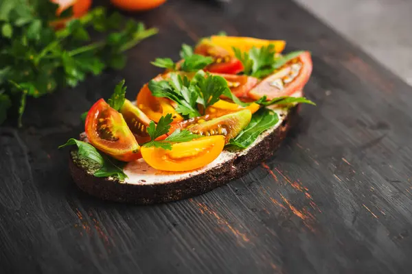 Healthy vegetarian sandwich with toasted bread, cheese, arugula, parsley and tomatoes on a dark wooden board.