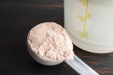 Plastik bir kaşık içindeki beyaz çikolatalı protein tozu koyu ahşap arka planda..
