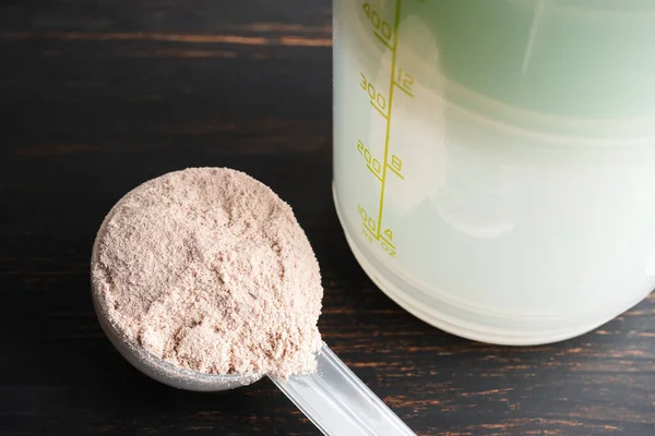 Whey chocolate protein powder in a plastic measuring spoon on a dark wooden background.