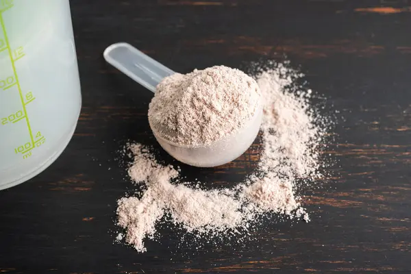 Whey chocolate protein powder in a plastic measuring spoon on a dark wooden background.