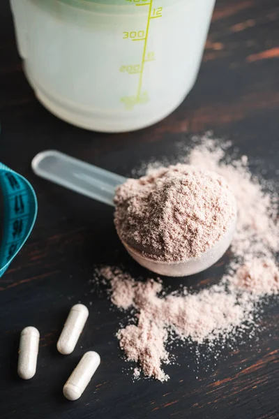 Scoop of chocolate whey protein isolate, white capsules of amino acids, creatine and measuring tape, bodybuilding food supplements on a dark wooden board.