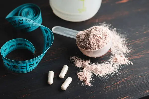 Scoop of chocolate whey protein isolate, white capsules of amino acids, creatine and measuring tape, bodybuilding food supplements on a dark wooden board.