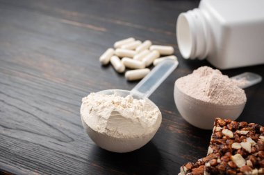 İki kepçe whey veya soya protein tozu, beyaz amino asit kapsülleri, vitaminler, kreatin, protein çikolatası, vücut geliştirme gıda malzemeleri, koyu ahşap bir tahtanın üzerinde spor besinleri..