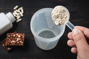 Erkek eli süt tozu kepçesinin içine soya proteini tozu koyar. Beyaz kapsüller amino asitler, vitaminler ve protein çikolataları. Koyu tahta tahtada, vücut geliştiren gıda takviyeleri..