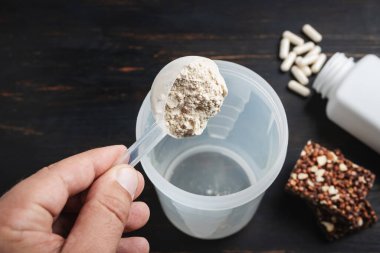 Erkek eli süt tozu kepçesinin içine soya proteini tozu koyar. Beyaz kapsüller amino asitler, vitaminler ve protein çikolataları. Koyu tahta tahtada, vücut geliştiren gıda takviyeleri..