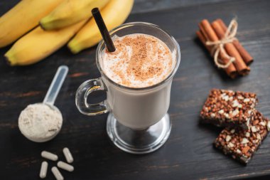 Bir bardak protein milkshake, saman ve bir kepçe beyaz protein tozu beyaz amino asit kapsülleri, vitaminler, protein barı ve muzlar koyu ahşap bir tahtanın üzerinde, vücut geliştiren gıda takviyeleri..