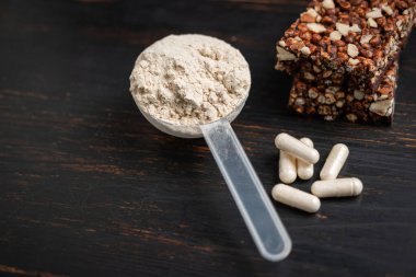 Bir kepçe whey veya soya protein tozu, beyaz amino asit kapsülleri, vitaminler, kreatin, protein çikolatası, vücut geliştirme gıdaları, koyu ahşap bir tahtanın üzerindeki spor gıdaları..