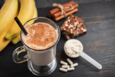 Bir bardak protein milkshake, saman ve bir kepçe beyaz protein tozu beyaz amino asit kapsülleri, vitaminler, protein barı ve muzlar koyu ahşap bir tahtanın üzerinde, vücut geliştiren gıda takviyeleri..