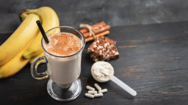 Bir bardak protein milkshake, saman ve bir kepçe beyaz protein tozu beyaz amino asit kapsülleri, vitaminler, protein barı ve muzlar koyu ahşap bir tahtanın üzerinde, vücut geliştiren gıda takviyeleri..