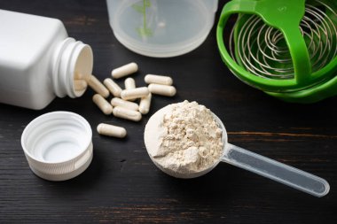 Süt veya soya proteini tozu, beyaz amino asit kapsülleri, vitaminler ve kreatin, vücut geliştiren gıda takviyeleri, spor beslenmesi, siyah tahta üzerinde plastik karıştırıcı..