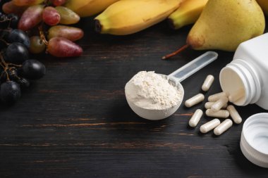 Bir kepçe süt veya soya protein tozu, beyaz amino asit kapsülleri, vitaminler, kreatin, vücut geliştiren gıda takviyeleri ve spor besinleri. Tahtada meyve, muz, armut ve üzüm.