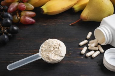 Bir kepçe süt veya soya protein tozu, beyaz amino asit kapsülleri, vitaminler, kreatin, vücut geliştiren gıda takviyeleri ve spor besinleri. Tahtada meyve, muz, armut ve üzüm.