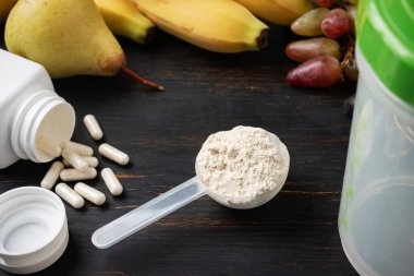 Bir kepçe süt veya soya protein tozu, beyaz amino asit kapsülleri, vitaminler, kreatin, vücut geliştiren gıda takviyeleri ve spor besinleri. Tahtada meyve, muz, armut ve üzüm.