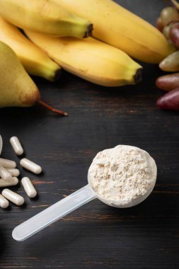 Bir kepçe süt veya soya protein tozu, beyaz amino asit kapsülleri, vitaminler, kreatin, vücut geliştiren gıda takviyeleri ve spor besinleri. Tahtada meyve, muz, armut ve üzüm.