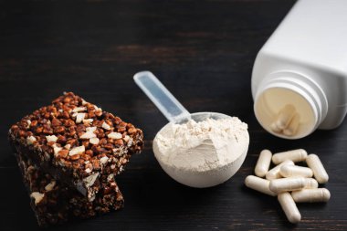 Bir kepçe whey veya soya protein tozu, beyaz amino asit kapsülleri, vitaminler, kreatin, protein çikolatası, vücut geliştirme gıdaları, koyu ahşap bir tahtanın üzerindeki spor gıdaları..