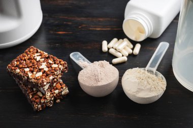 İki kepçe whey veya soya protein tozu, beyaz amino asit kapsülleri, vitaminler, kreatin, protein çikolatası, vücut geliştirme gıda malzemeleri, koyu ahşap bir tahtanın üzerinde spor besinleri..