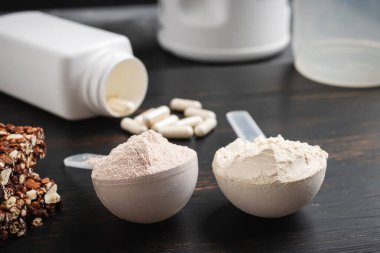 İki kepçe whey veya soya protein tozu, beyaz amino asit kapsülleri, vitaminler, kreatin, protein çikolatası, vücut geliştirme gıda malzemeleri, koyu ahşap bir tahtanın üzerinde spor besinleri..