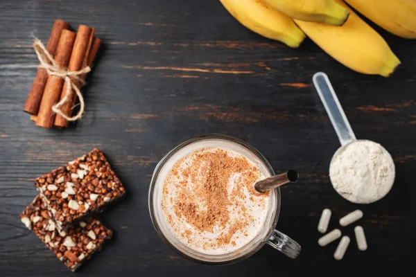 Glass of protein milkshake drink with straw and scoop of whey protein powder, white capsules of amino acids, vitamins, protein bar and bananas on a dark wooden board, top view.