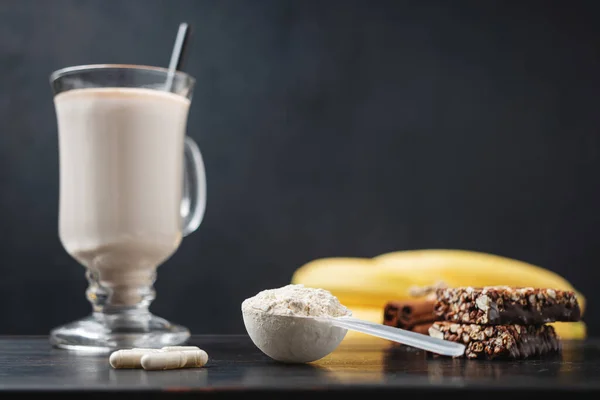 Scoop of whey protein powder and glass of protein milkshake drink with straw, white capsules of amino acids, vitamins, protein bar on a dark wooden board, bodybuilding food supplements.