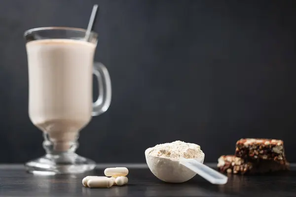 Scoop of whey protein powder and glass of protein milkshake drink with straw, white capsules of amino acids, vitamins, protein bar on a dark wooden board, bodybuilding food supplements.