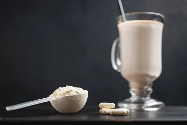 Scoop of whey protein powder and glass of protein milkshake drink with straw, white capsules of amino acids on a dark wooden board, bodybuilding food supplements.