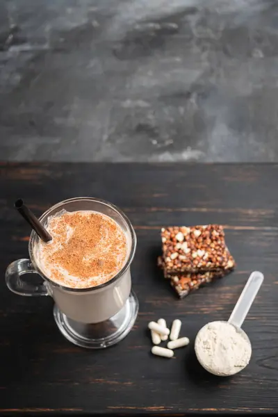 Glass of protein milkshake drink with straw and scoop of whey protein powder, white capsules of amino acids, vitamins, protein bar on a dark wooden board, bodybuilding food supplements.