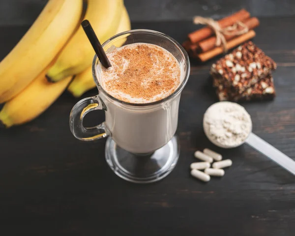 Glass of protein milkshake drink with straw and scoop of whey protein powder, white capsules of amino acids, vitamins, protein bar and bananas on a dark wooden board, bodybuilding food supplements.