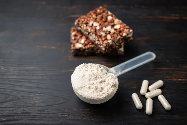 Scoop of whey or soy protein powder, white capsules of amino acids, vitamins, creatine, protein chocolate bar, bodybuilding food supplements, sports nutrition on a dark wooden board.