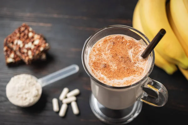 Glass of protein milkshake drink with straw and scoop of whey protein powder, white capsules of amino acids, vitamins, protein bar and bananas on a dark wooden board, bodybuilding food supplements.