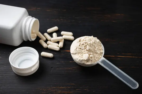 Scoop of whey or soy protein powder, white capsules of amino acids, vitamins and creatine, bodybuilding food supplements, sports nutrition on a dark wooden board.