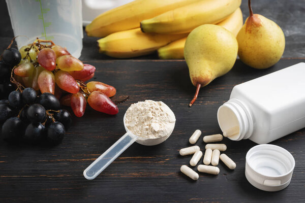 Scoop of whey or soy protein powder, white capsules of amino acids, vitamins, creatine, bodybuilding food supplements and sports nutrition. fruits, banana, pear and grape on a dark wooden board.