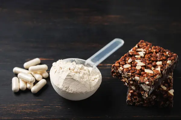 Scoop of whey or soy protein powder, white capsules of amino acids, vitamins, creatine, protein chocolate bar, bodybuilding food supplements, sports nutrition on a dark wooden board.