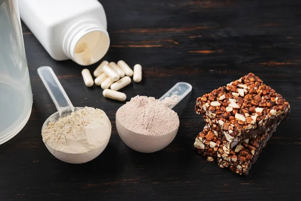 Two scoops of whey or soy protein powder, white capsules of amino acids, vitamins, creatine, protein chocolate bar, bodybuilding food supplements, sports nutrition on a dark wooden board.