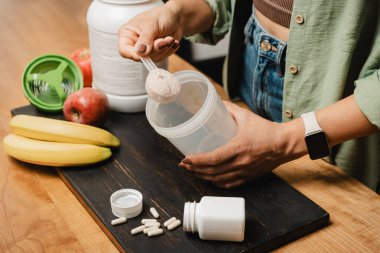 Elinde kaşık olan kot pantolonlu ve gömlekli bir kadın beyaz kapsüllü, muzlu ve elmalı beyaz protein tozunun porsiyonunu bir karıştırıcının içine koyar. Protein içeceği yapma süreci.