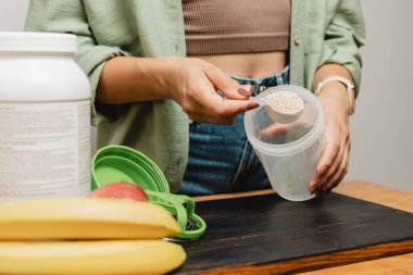 Elinde kaşık olan kot pantolonlu ve gömlekli bir kadın beyaz kapsüllü, muzlu ve elmalı beyaz protein tozunun porsiyonunu bir karıştırıcının içine koyar. Protein içeceği yapma süreci.